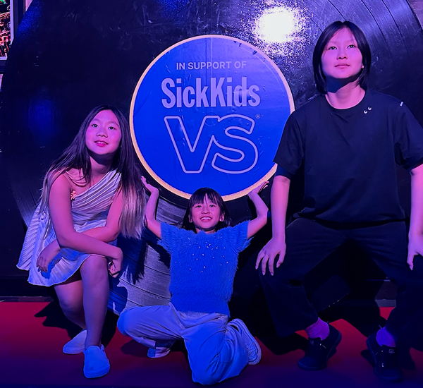 Alt text for Mad Carrot: Charlotte, Olivia and Rebecca, members of the Mad Carrot, posing with a sign that has the SickKids logo on it.  