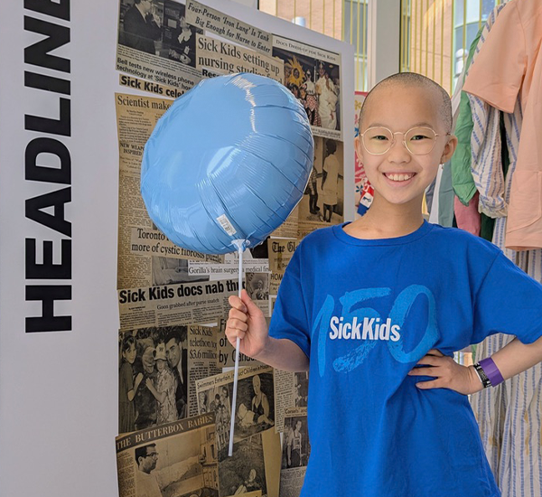 Nori smiling, with her hand on her hip, holding a blue balloon and wearing a SickKids t-shirt. 