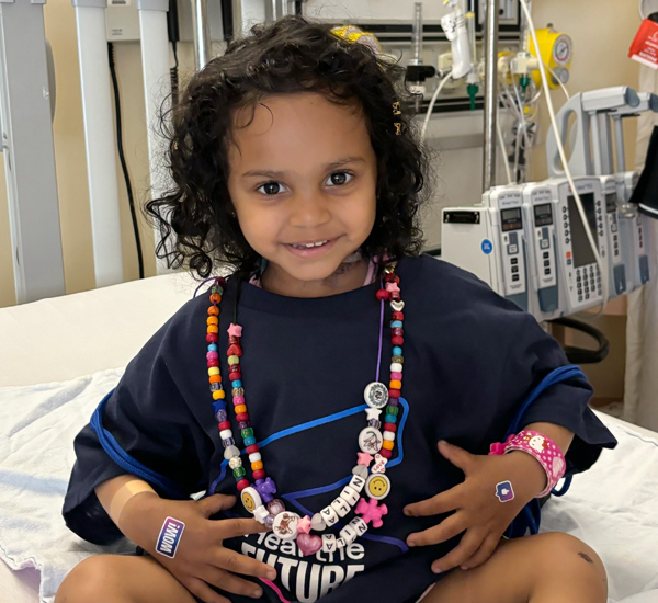 Nila sitting on her bed at SickKids. She’s smiling and wearing bravery beads. 