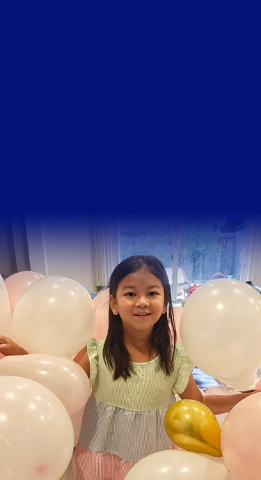Thea at home, smiling and surrounded by pink and gold balloons.