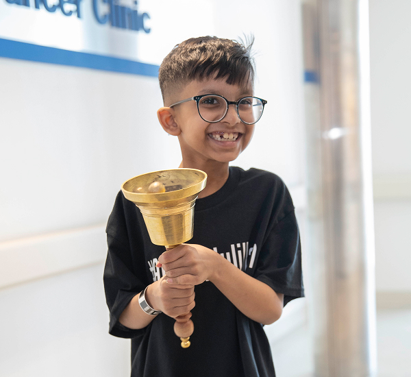 Qais at SickKids, smiling and holding a golden bell.