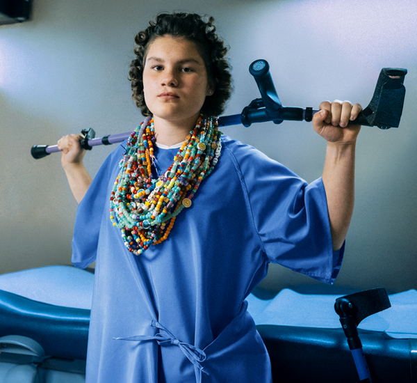 Wynn at SickKids. She’s wearing many strands of colourful bravery beads and holding one of her crutches over her head.