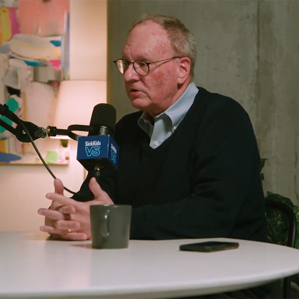 Dr. Jim Whitlock, wearing a light blue button-down shirt with a black sweater overtop, is seated in a creative studio and captured mid-conversation. A multi-coloured painting hangs behind him with a microphone in front of him. 