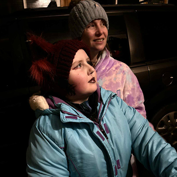 Nika (front) wears a light blue winter coat and is adorned with Hallowe'en makeup. She stands outdoors with her mom, Amanda, behind her, who is in a multi-coloured tie-dye sweater and grey beanie. Both are looking toward something off-camera..