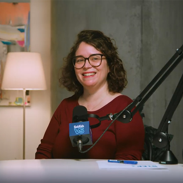 Dr. Sarah Cohen-Gogo is wearing a deep red sweater and is seated in a creative studio. She is looking off-camera and smiling. A multi-coloured painting hangs behind her, along with a soft-lit lamp. A microphone is perched in front of her.