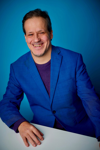A professional headshot of Jon who is smiling at the camera. He wears a blue blazer and shirt and sits behind a table in front of a blue background.