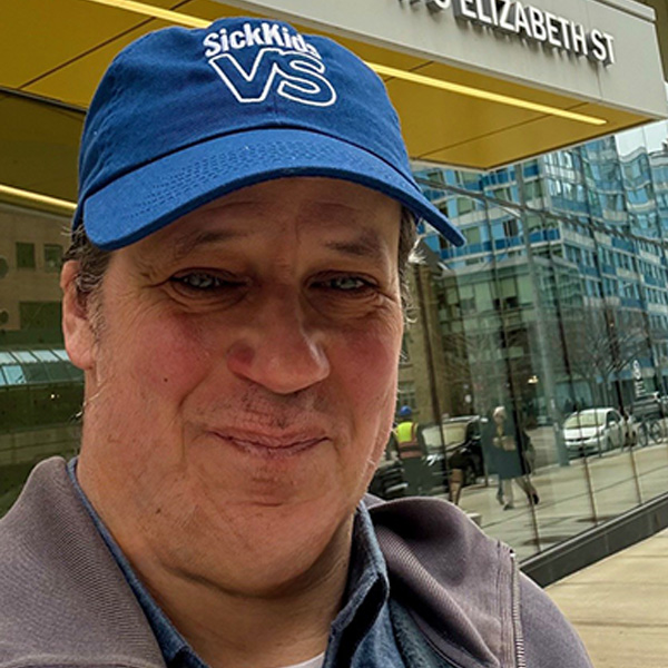 A selfie of Jon standing in front of the entrance to the SickKids Patient Support Centre. He is wearing a blue SickKidsVS baseball cap, grey sweater, and blue collared shirt. 