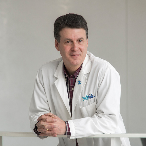 Dr. Stephen Scherer, SickKids Chief of Research, stands leaning over a bannister. He has short, dark hair and is wearing a white lab coat with SickKids embroidered on it over a collared shirt.