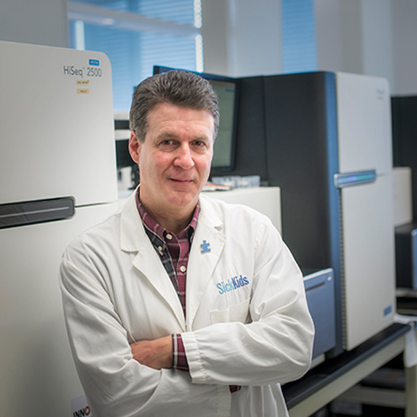 Dr. Stephen Scherer, SickKids Chief of Research, stands in a laboratory at SickKids. He has short, dark hair and is wearing a white lab coat with SickKids embroidered on it over a collared shirt.