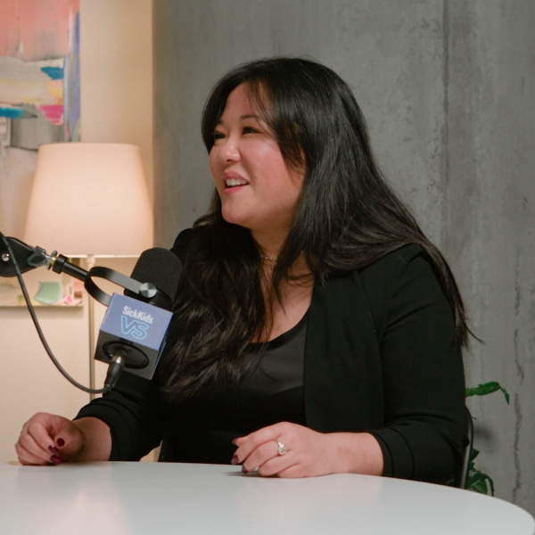 Anna, a genetics counsellor at SickKids, is seated in a creative studio with multi-coloured paintings on the wall, white walls, potted plants, and a table with chairs and mics. She speaks with the SickKids VS Podcast host, Hannah Bank. Anna is wearing a black blazer with a skirt and her long, black hair flowing past her shoulders. Hannah is wearing a black top with pants and her dark brown, curly hair flowing past her shoulders.