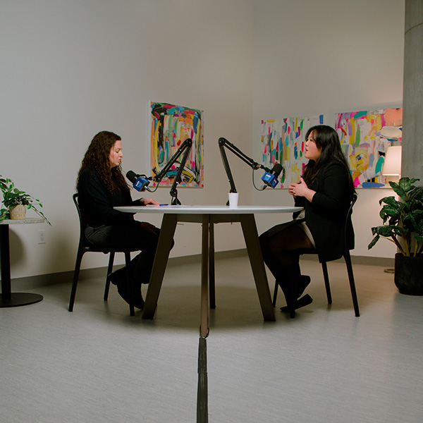 Anna, a genetics counsellor at SickKids, is seated in a creative studio with multi-coloured paintings on the wall, white walls, potted plants, and a table with chairs and mics. She speaks with the SickKids VS Podcast host, Hannah Bank. Anna is wearing a black blazer with a skirt and her long, black hair flowing past her shoulders. Hannah is wearing a black top with pants and her dark brown, curly hair flowing past her shoulders.