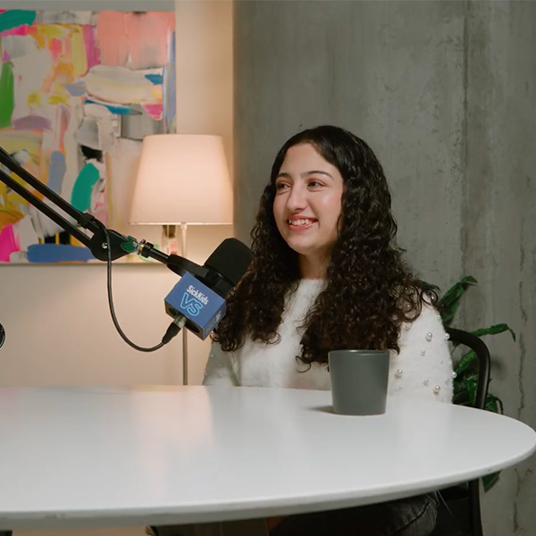 Melika, a former SickKids patient, is seated in a creative studio with multi-coloured paintings on the wall, white walls, potted plants, and a table with chairs and mics. She speaks with the SickKids VS Podcast host, Hannah Bank. Both women are wearing white sweaters, jeans, and have long, curly, dark hair.